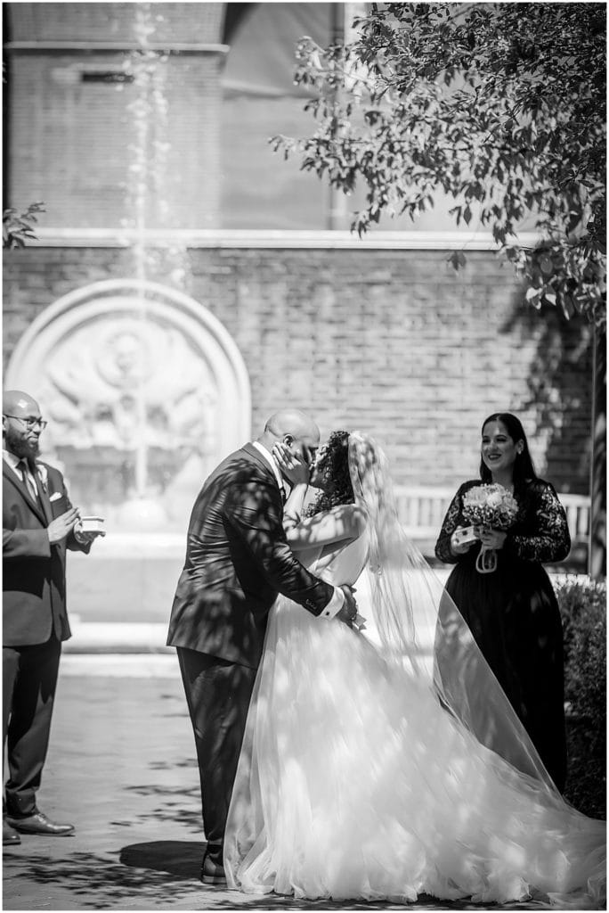 Black and white portrait of bride and groom kissing during intimate outdoor micro wedding ceremony in Penn Museum garden courtyard