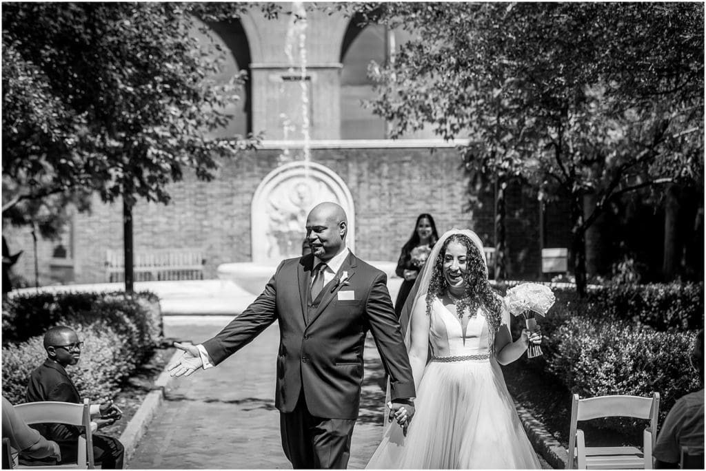 Black and white portrait of bride and groom walking up aisle in garden courtyard of Penn Museum after intimate micro wedding ceremony