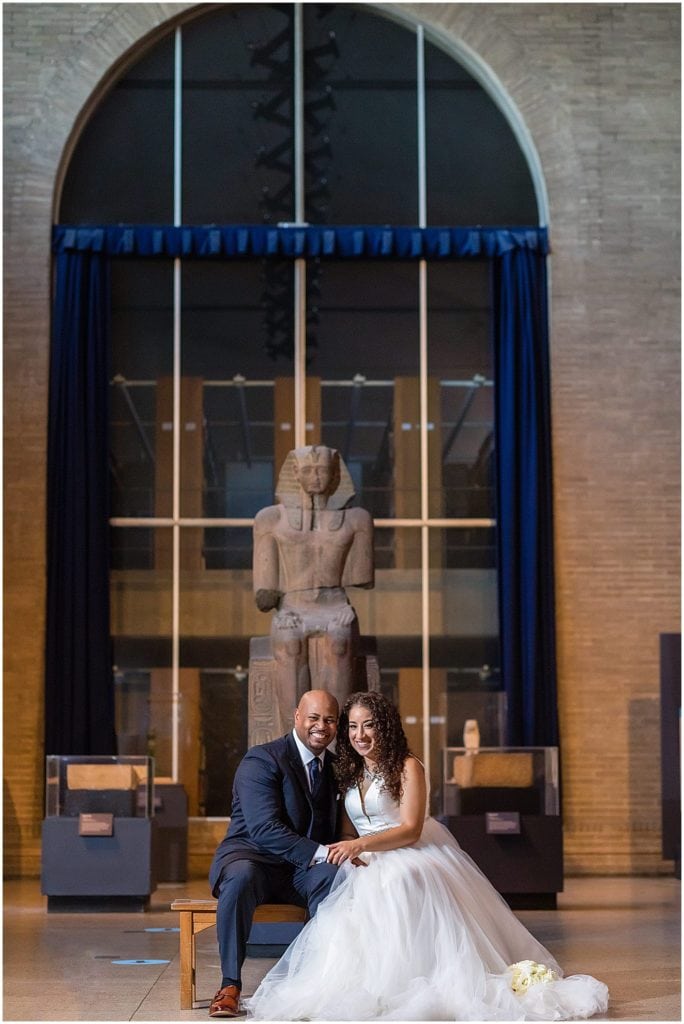 Bride and groom sit on a bench in front of Egyptian artifacts in Penn Museum micro wedding