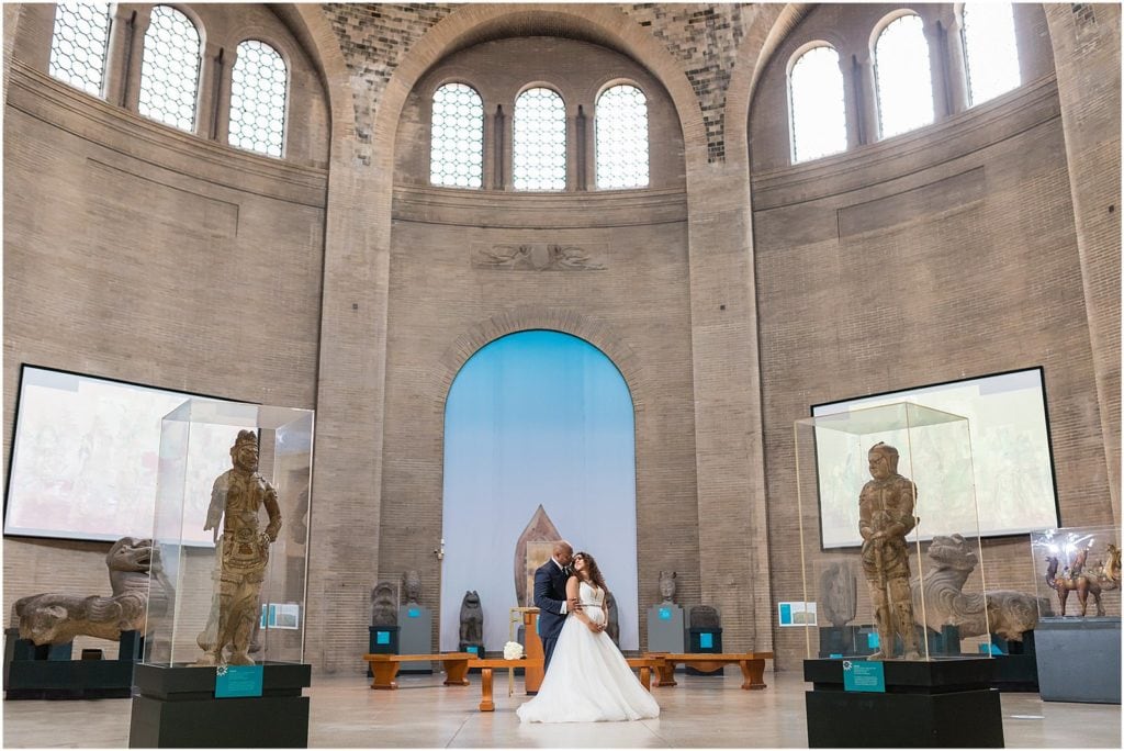 Bride and groom lean in for a kiss in the center of statue exhibit in Penn Museum micro wedding