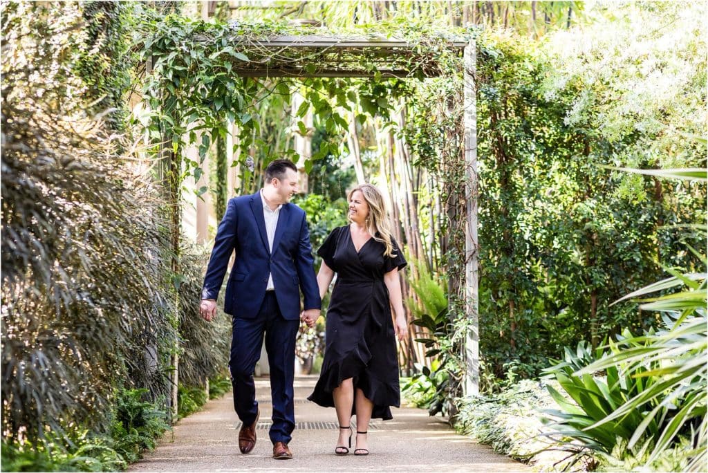 Bride and groom walk holding hands over greenery covered bridge at Longwood Gardens engagement session