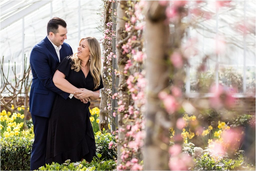 Couple hugging and laughing next to floral wall in Longwood Gardens greenhouse engagement session