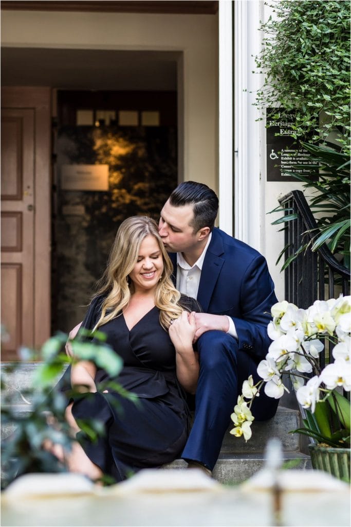 Groom kissing bride on forehead while sitting on steps in front of Longwood Gardens door during engagement session