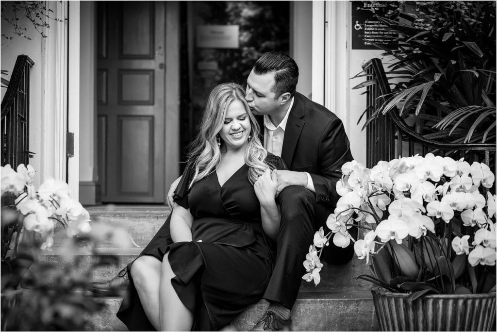 Black and white portrait of groom kissing bride on forehead while sitting on steps at Longwood Gardens romantic engagement session