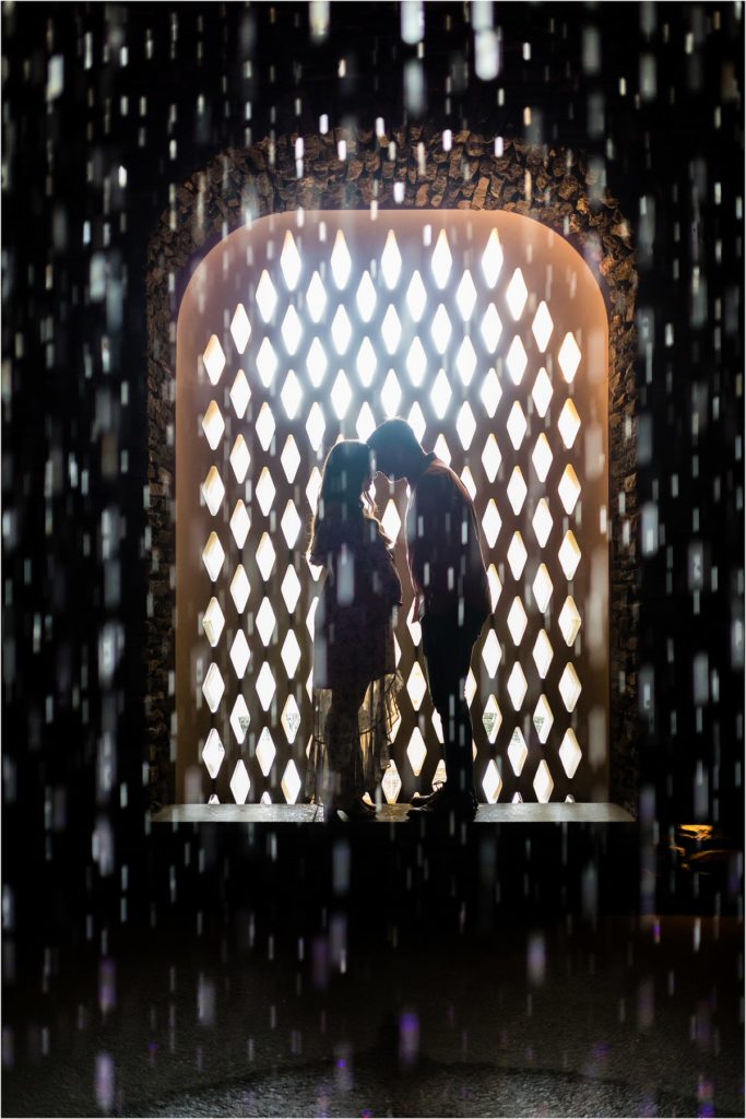 Husband and wife lean in for kiss behind rain fountain at Longwood Gardens maternity session