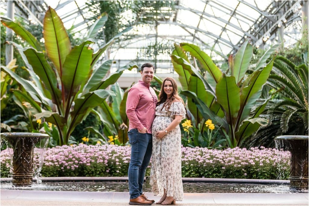 Husband and wife stand in front of fountain in greenhouse at Longwood Gardens maternity session