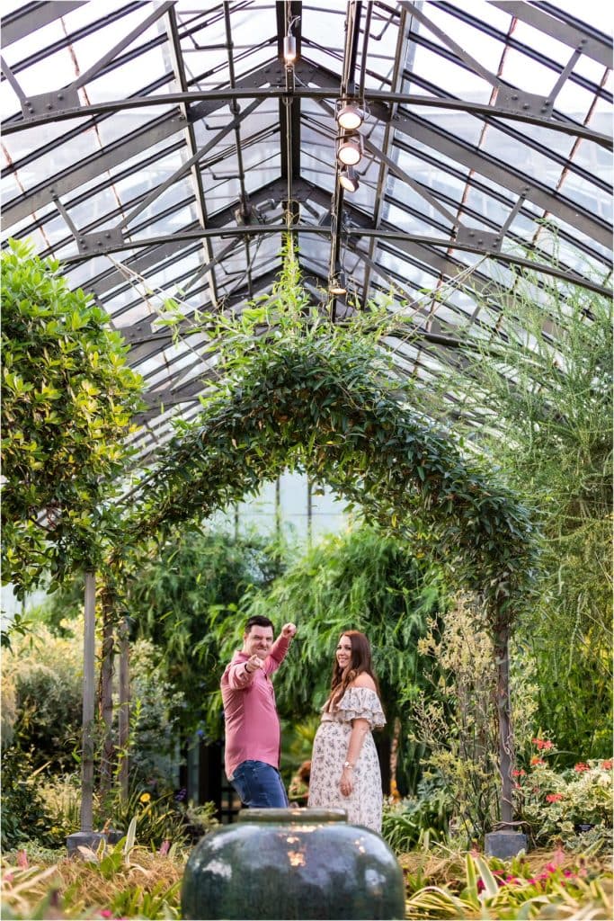 Husband dancing and pointing to camera with wife laughing during maternity shoot in Longwood Gardens greenhouse