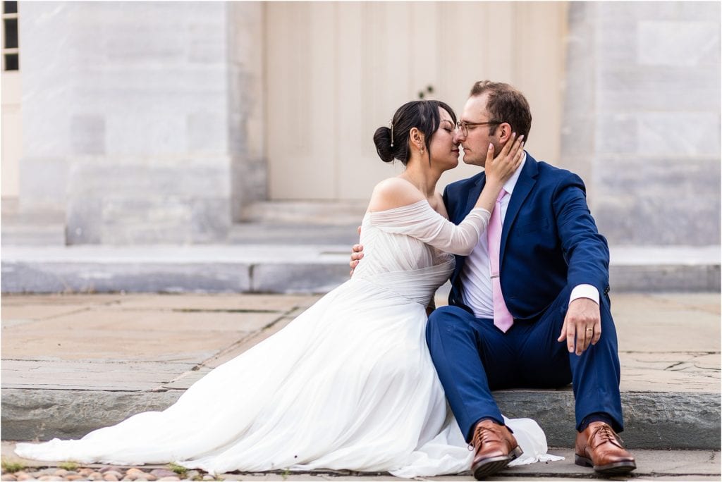 Bride pulls groom in for a kiss while they sit on a ledge at the Merchant Exchange Building in Old City Philadelphia