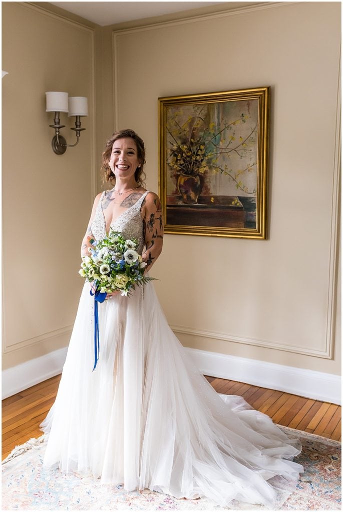 Window lit portrait of bride holding blue and white bouquet at Bolingbroke Mansion same sex wedding
