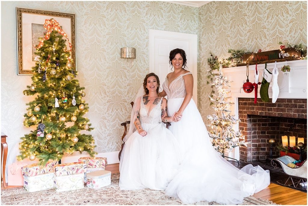 Traditional same sex marriage portrait of brides sitting next to Christmas tree and decorated fireplace in September same sex wedding
