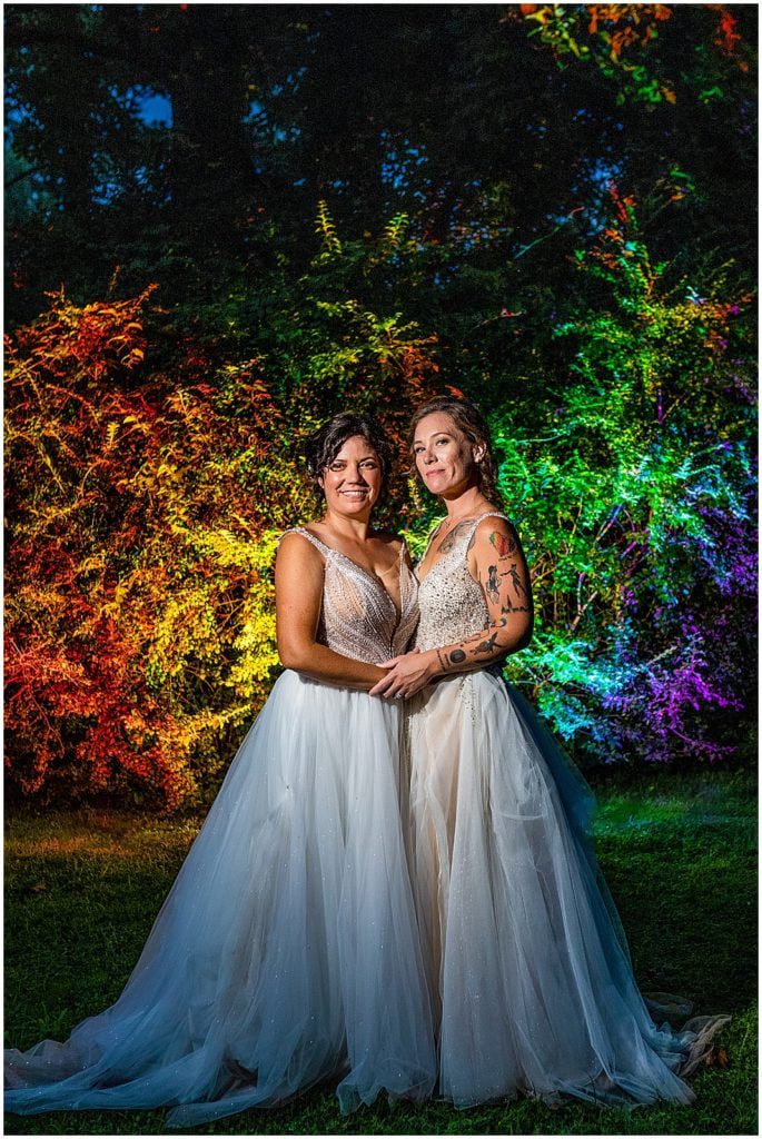 Brides stand together in front of rainbow lit trees at same sex Pride wedding at Bolingbroke Mansion
