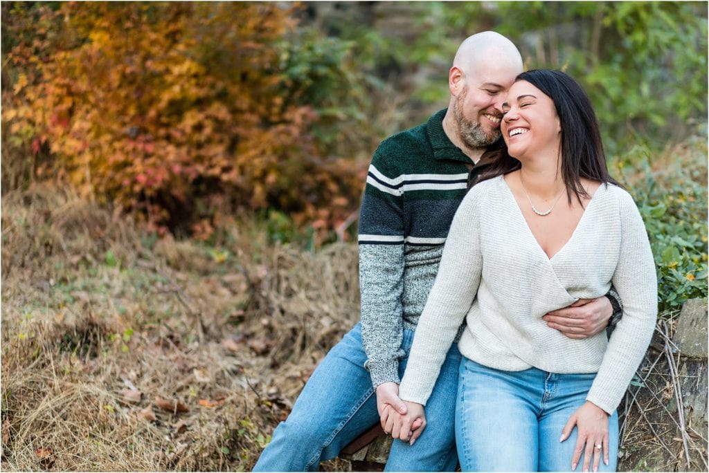 Man holding fiancé on his lap sitting on stone wall at Ridley Creek Park engagement session