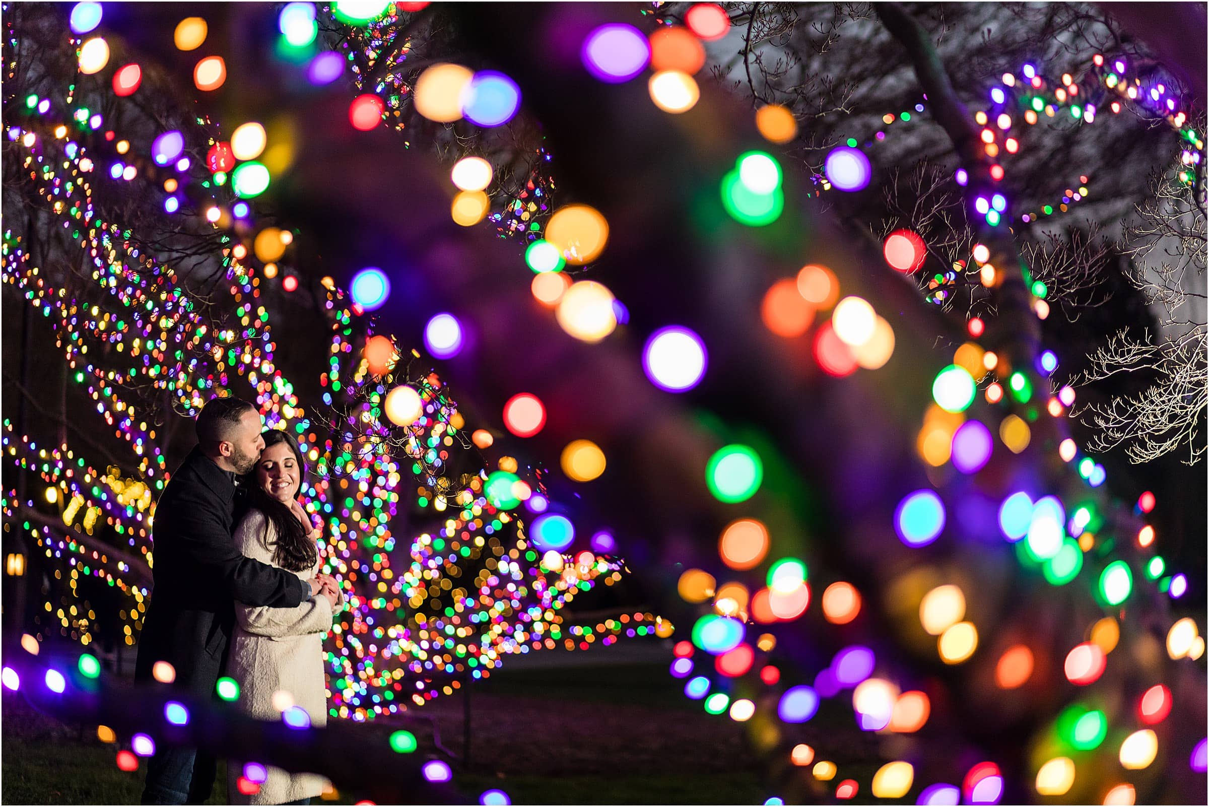 Man kissing his fiancée on the forehead through colorful light wrapped trees at Longwood Gardens Christmas engagement session