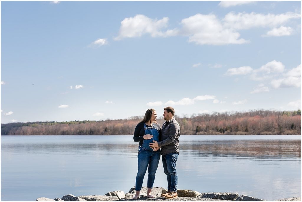 Husband and wife stand on rock ledge in front of lake holding belly in Pocono Mountain maternity session portrait