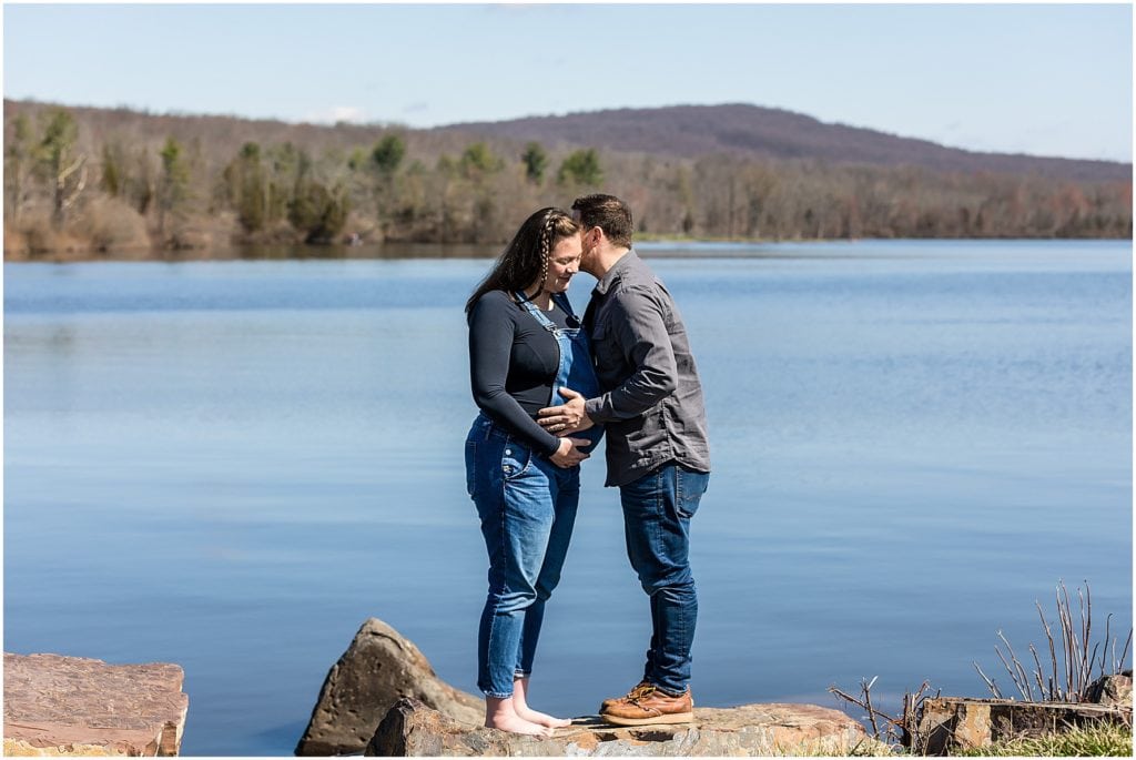 Husband holding wife's belly while whispering in her ear in front of lake at Pocono Mountains maternity couple session