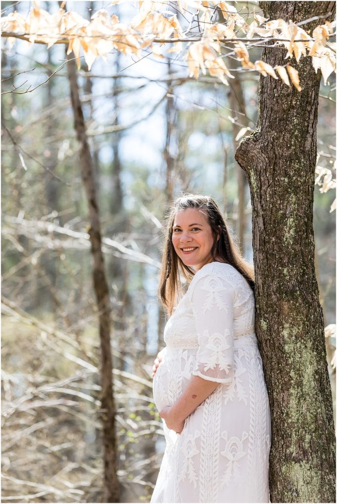 Expecting mother in white lace dress leans against tree while holding her baby belly in Pocono Mountain maternity session