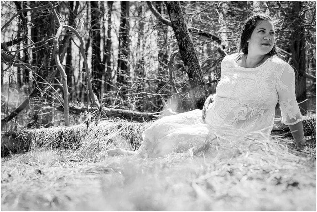 Black and white maternity session portrait of mother in white lace dress sitting in wooded grass