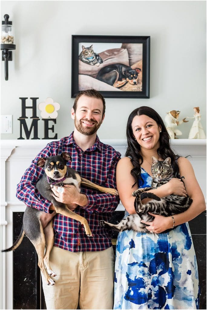 Couple smiling while holding their dog and cat standing in front of their fireplace at their home in Philadelphia