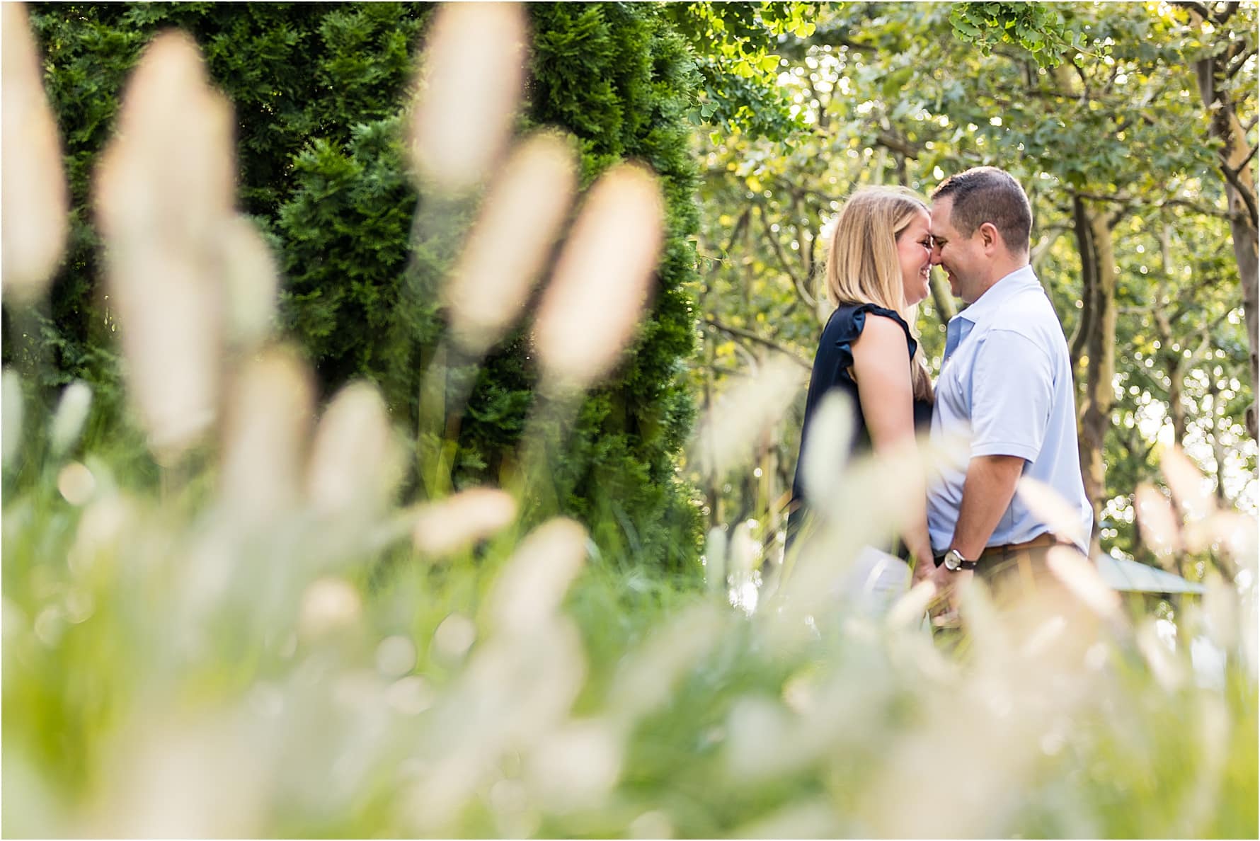 Bride and groom holding hands and smiling at one another touching foreheads as they stand in greenery and flowers