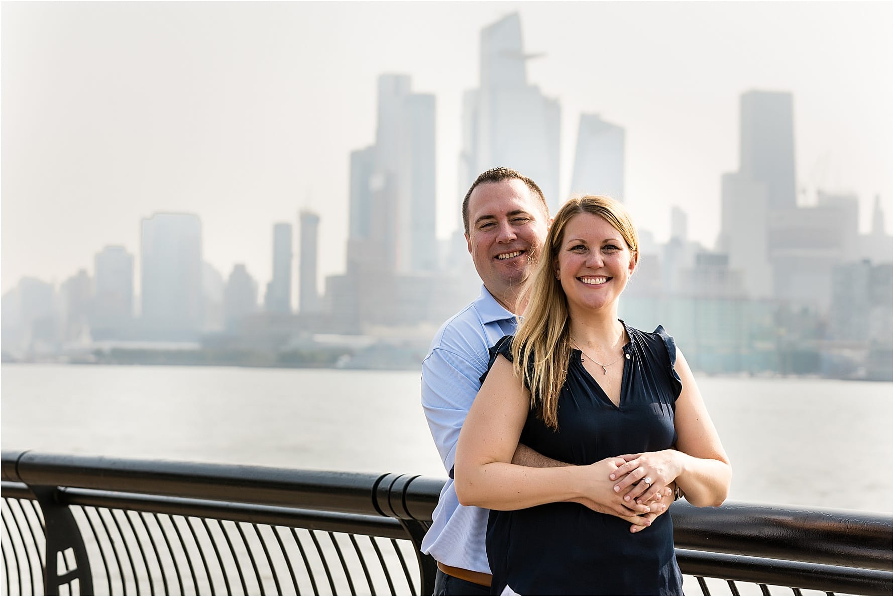 Groom holding the bride and smiling at the camera as they lean against a railing with the New York City skyline in the background