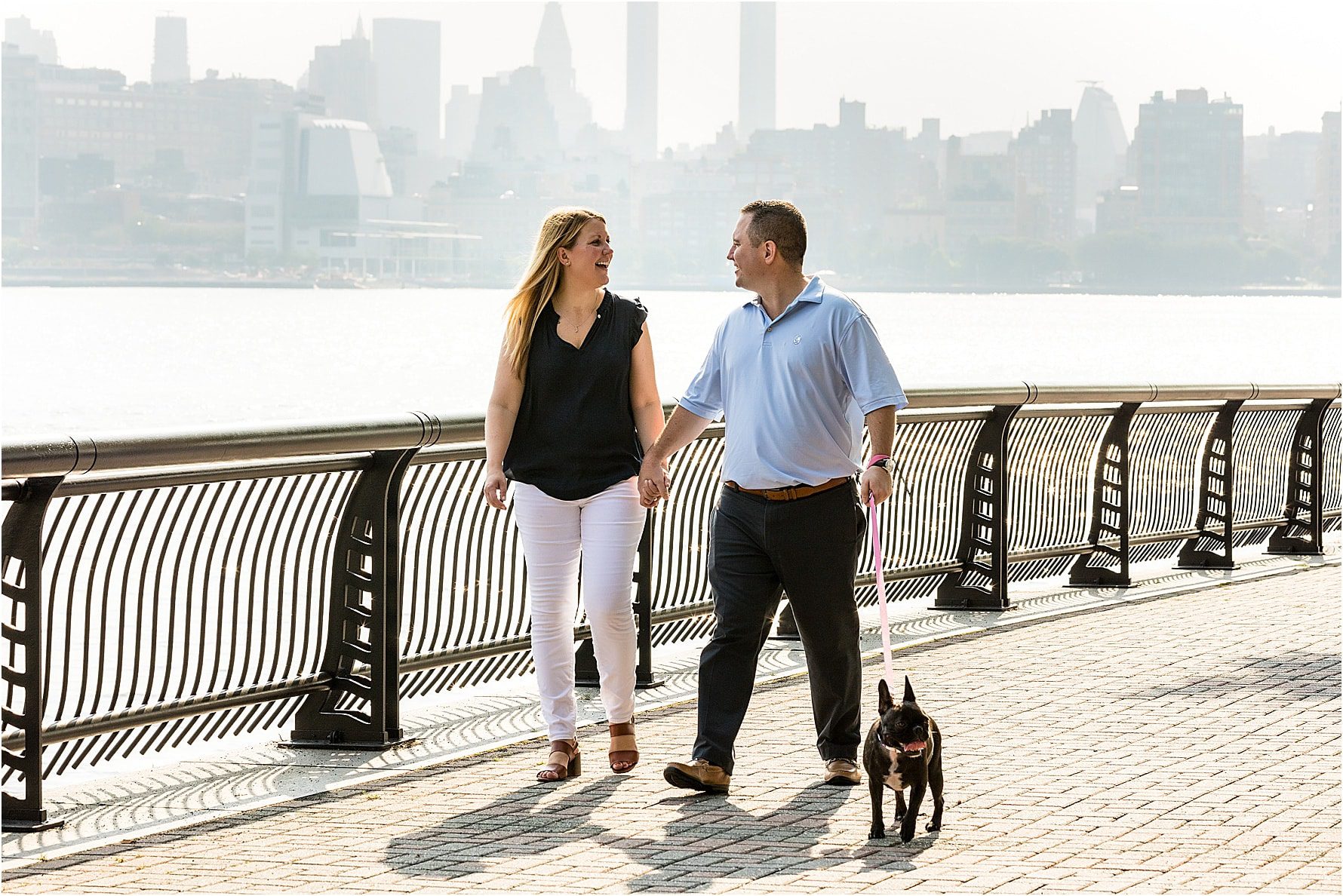 Bride and groom holding hands and smiling at each other as they walk their dog along the path with the New York City skyline behind them