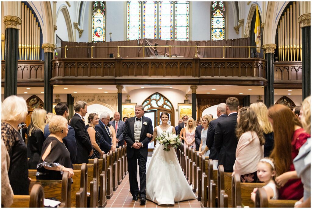 Bride walks down the aisle towards her groom accompanied by her father 