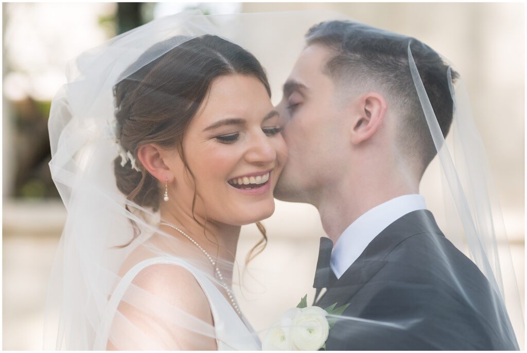 Bride smiles as her Groom kisses her cheek as her veil softly covers them