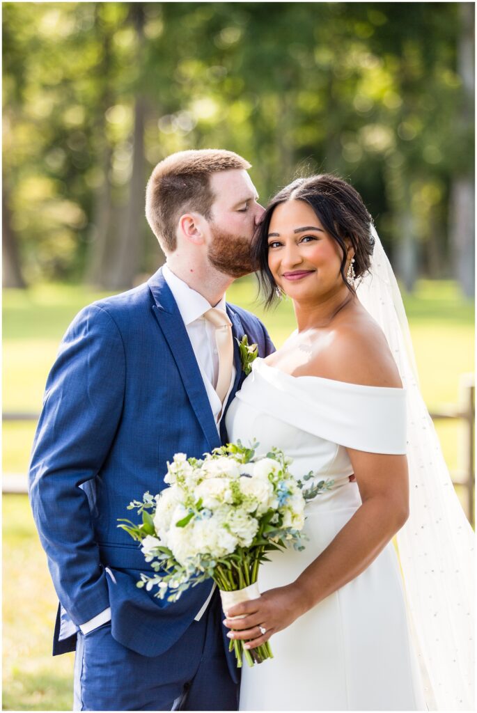 Groom kisses his bride as she holds her bouquet and smiles at the camera