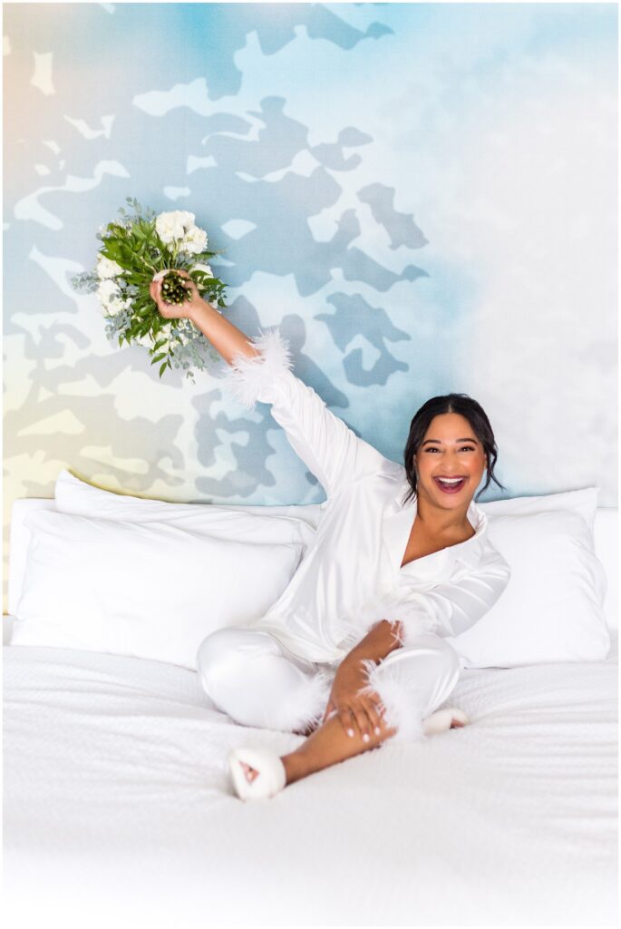 Bride smiles while holding her bouquet up high with one hand as she sits on a bed in a white outfit