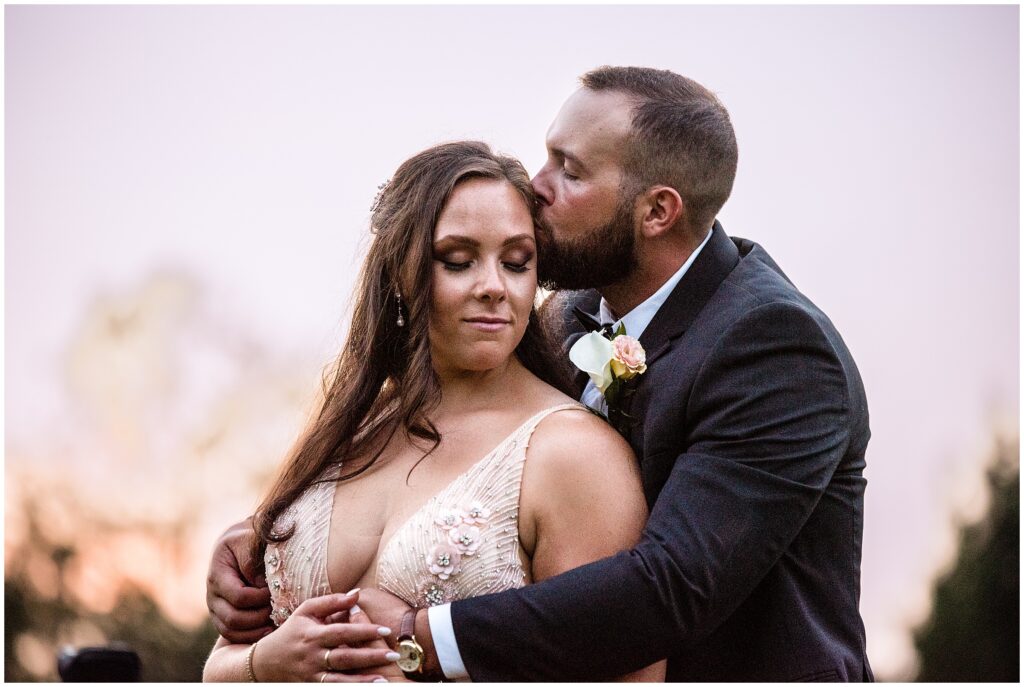 Groom kisses his bride as they embrace at sunset