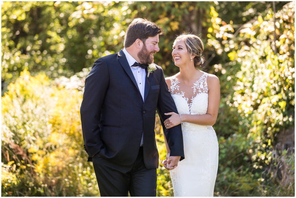 Groom and bride smile at each other and hold hands while walking on grounds at Loch Aerie Mansion
