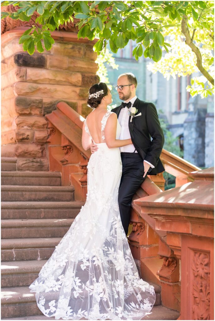 Groom stands with his Bride on stairs. Her wedding dress has a low back and gorgeous lace appliqués