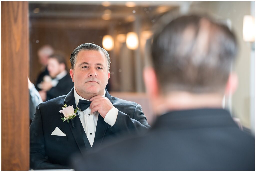 Groom adjusts his black bow tie in a mirror before wedding