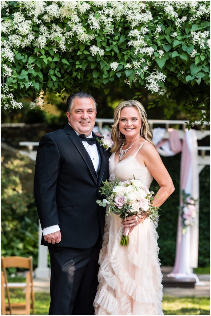 Groom in black tux and bow tie with his Bride, holding her pink and white rose bouquet in a pale pink ruffled wedding dress