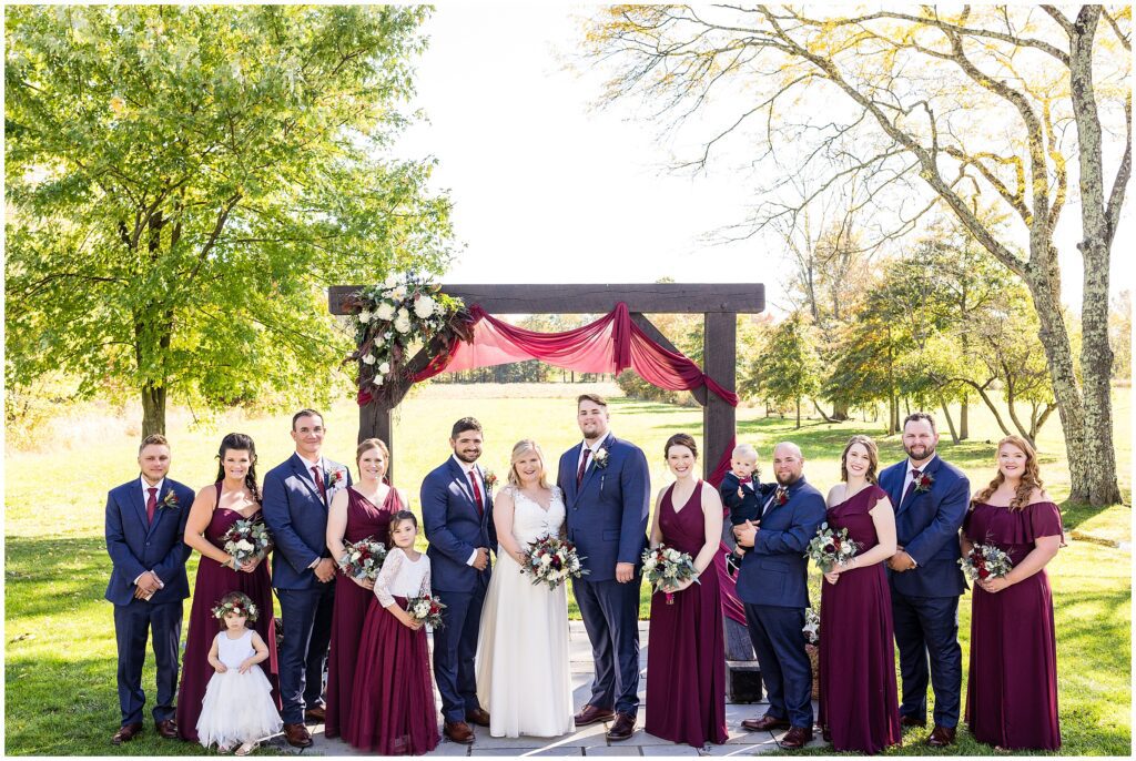 Wedding party in shades of deep red and navy near ceremony arch for fall wedding