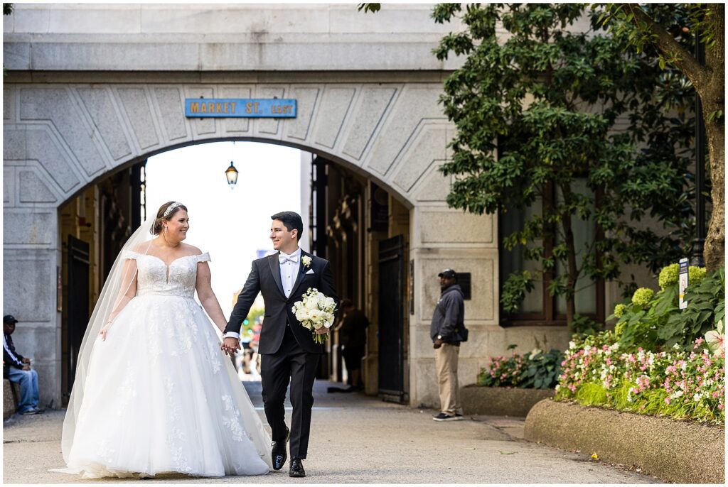 Bride and Groom hold hands while looking at each other and smiling, and groom holds the bride's white floral bouquet 