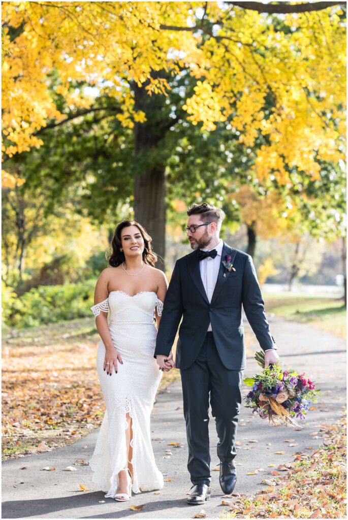 couple walks together holding hands, the sidewalk is surrounded by fallen autumn leaves and the trees behind them have leaves that are turning yellow