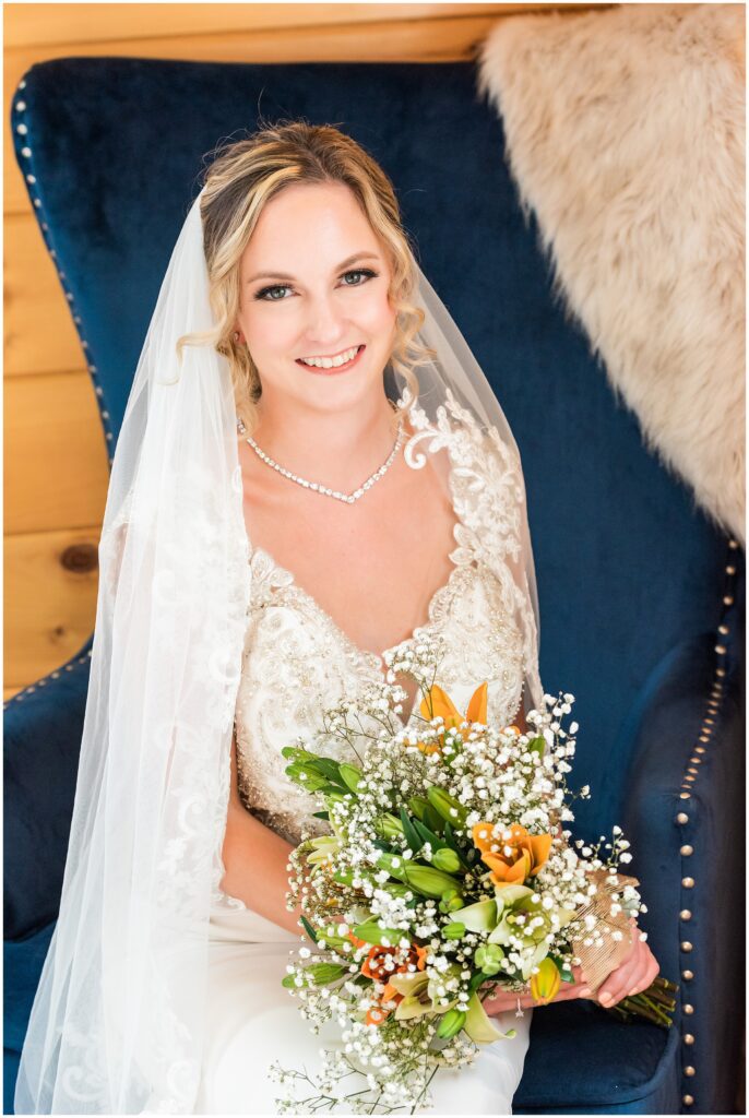 portrait of bride sitting in blue chair wearing veil, lace detail wedding dress, and holding bouquet