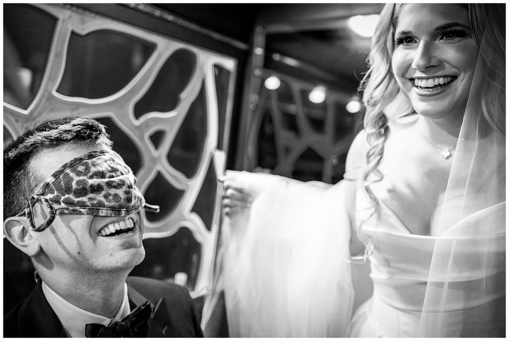 Black and white portrait of a blindfolded groom smiling as bride walks by him on the party bus before their first look | Ashley Gerrity Photography