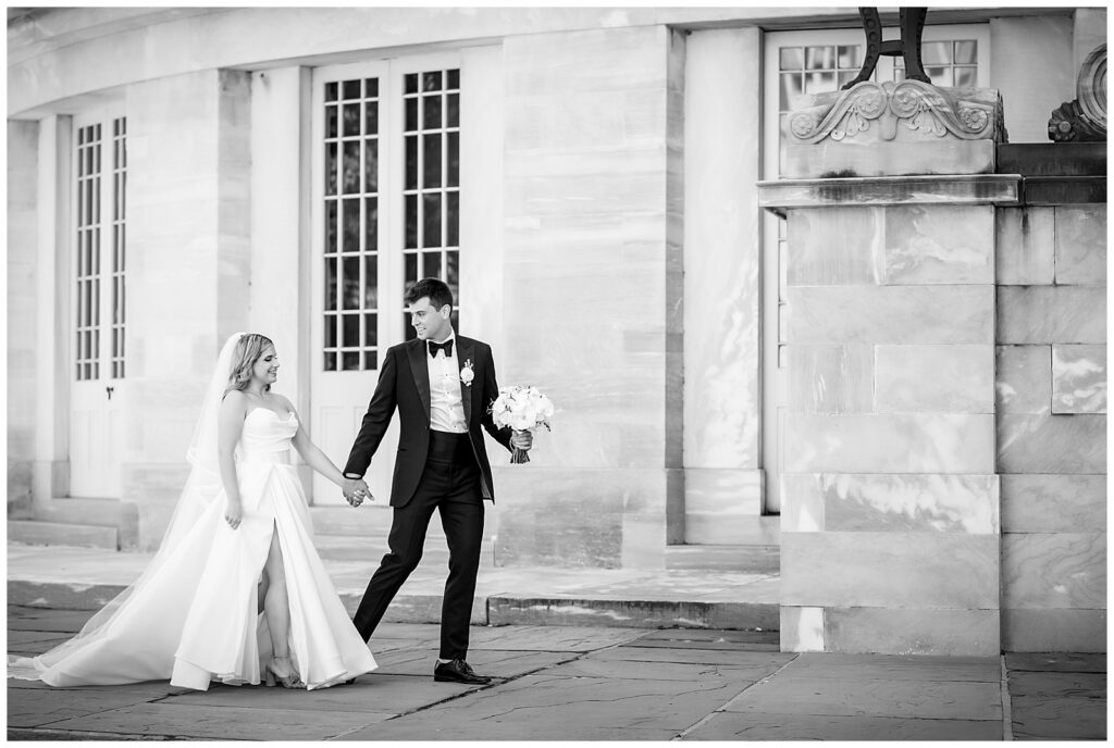 Black and white wedding portrait of groom holding bride flowers in one hand and brides hand in the other while walking through Old City Philadelphia | Ashley Gerrity Photography