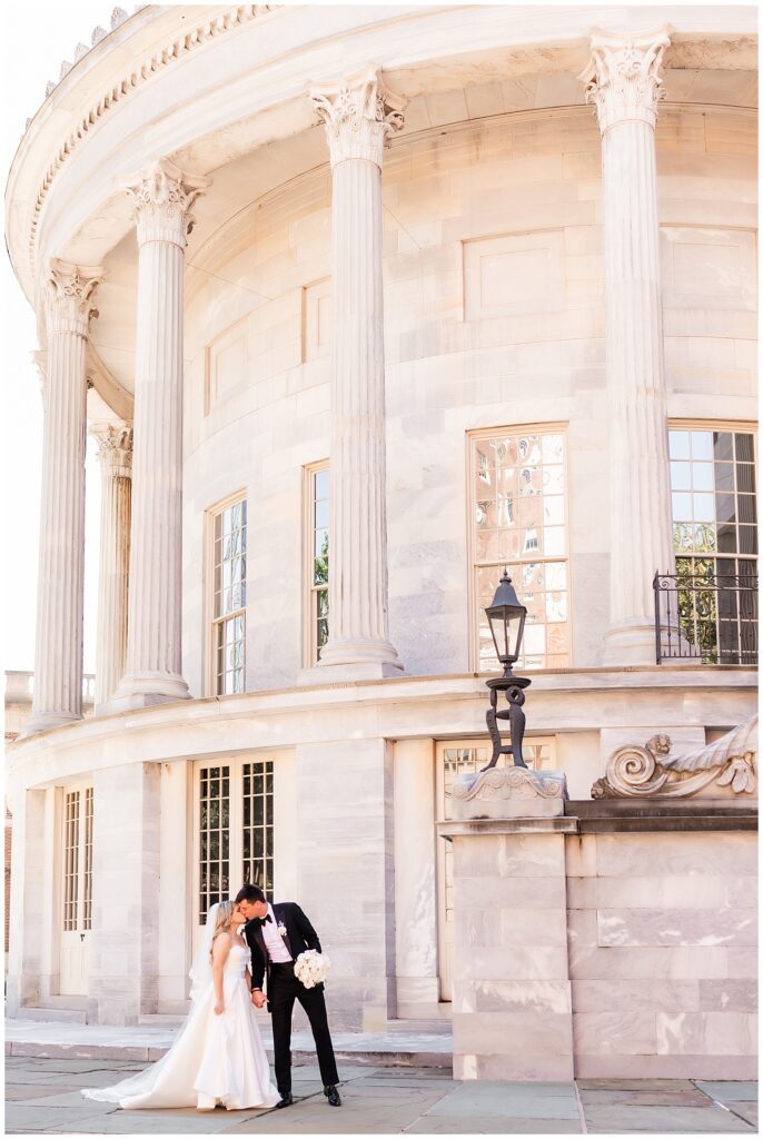 Bride and groom hold hands and kiss in front of tall marble building with columns in Old City Philadelphia | Ashley Gerrity Photography