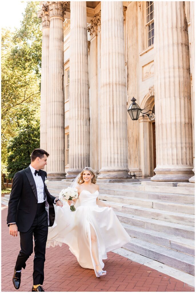 Groom holding brides a-line dress train as they walk in front of marble building with columns in Old City Philadelphia | Ashley Gerrity Photography