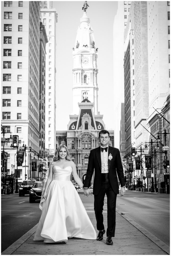 Black and white wedding portrait of bride and groom holding hands and walking down Broad Street in front of Philadelphia City Hall | Ashley Gerrity Photography