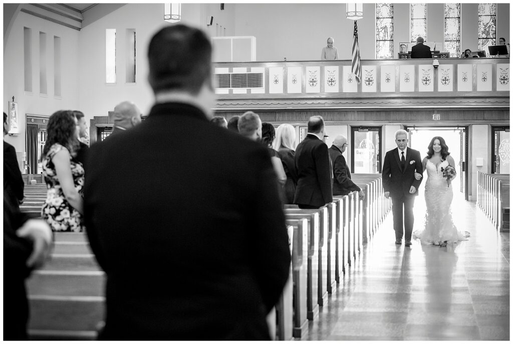 Black and white portrait of bride and her father walking down the aisle with groom in the foreground watching | Ashley Gerrity Photography
