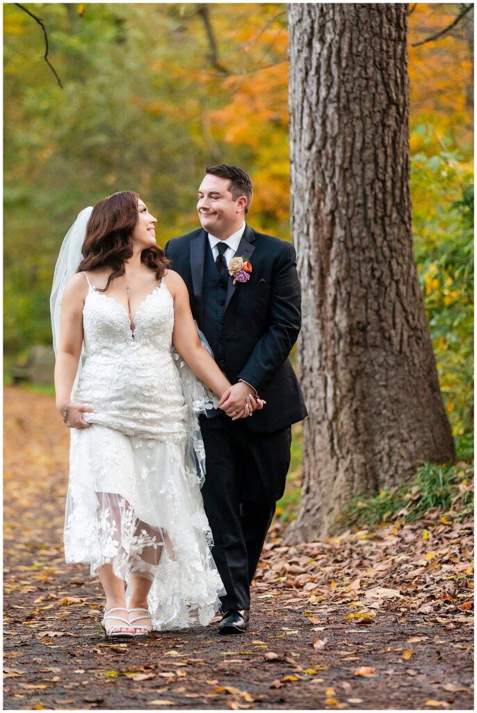 Bride holding her lace dress train while walking on park path during Fall Pennypack Park wedding portraits | Ashley Gerrity Photography