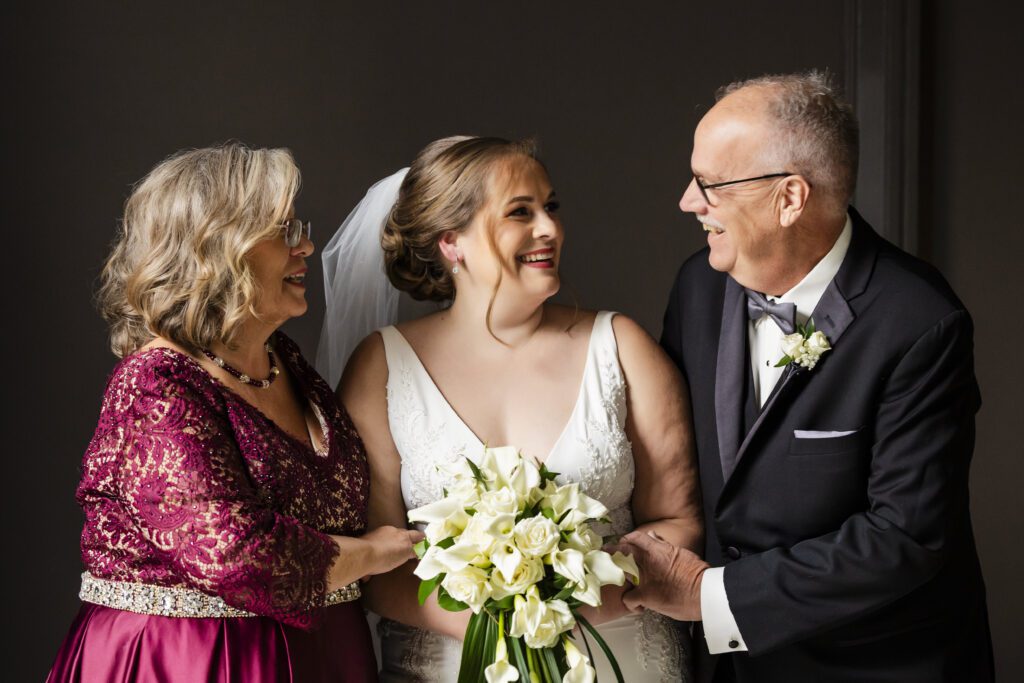 Bride in between her parents smiling against a dark background