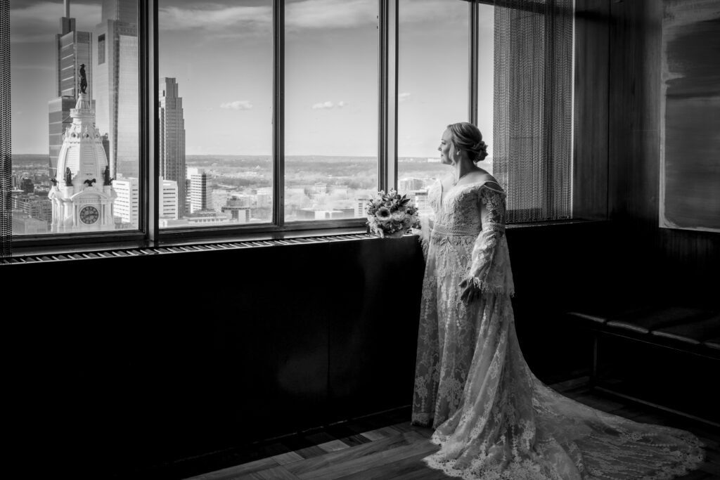 Black & white photo of a bride looking out the window of the 33rd floor of the Loews Philadelphia at the skyline
