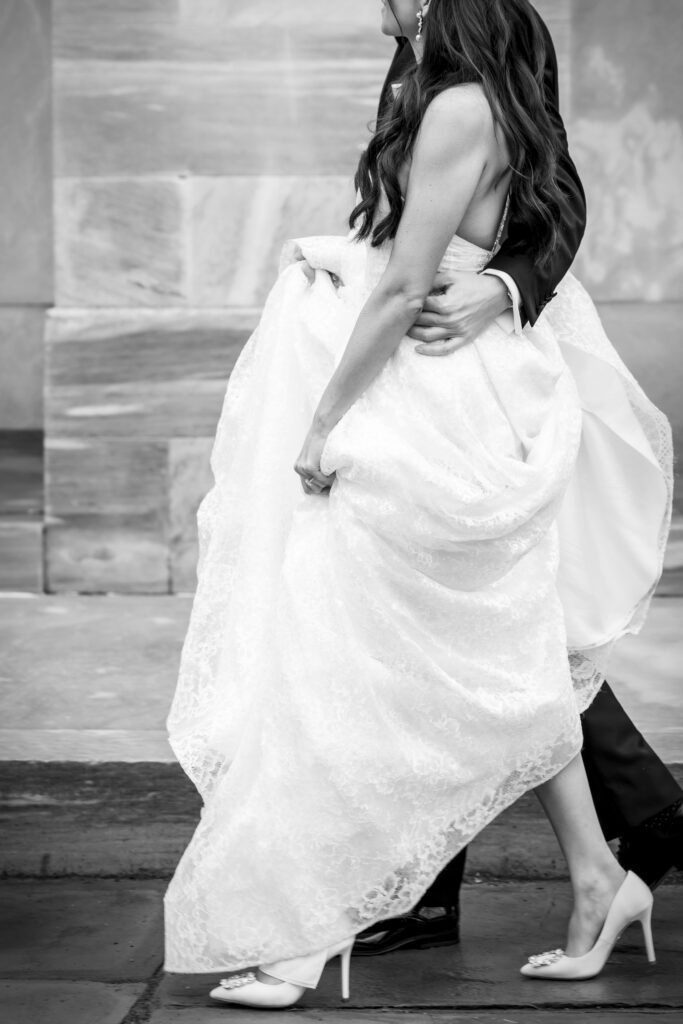 Black & white photo of bride holding up her dress as she walks, exposing high stilleto heels
