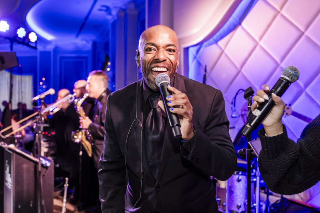 African American man in a suit with blue nail polish sings into a microphone while looking directly at the camera with band members in the background