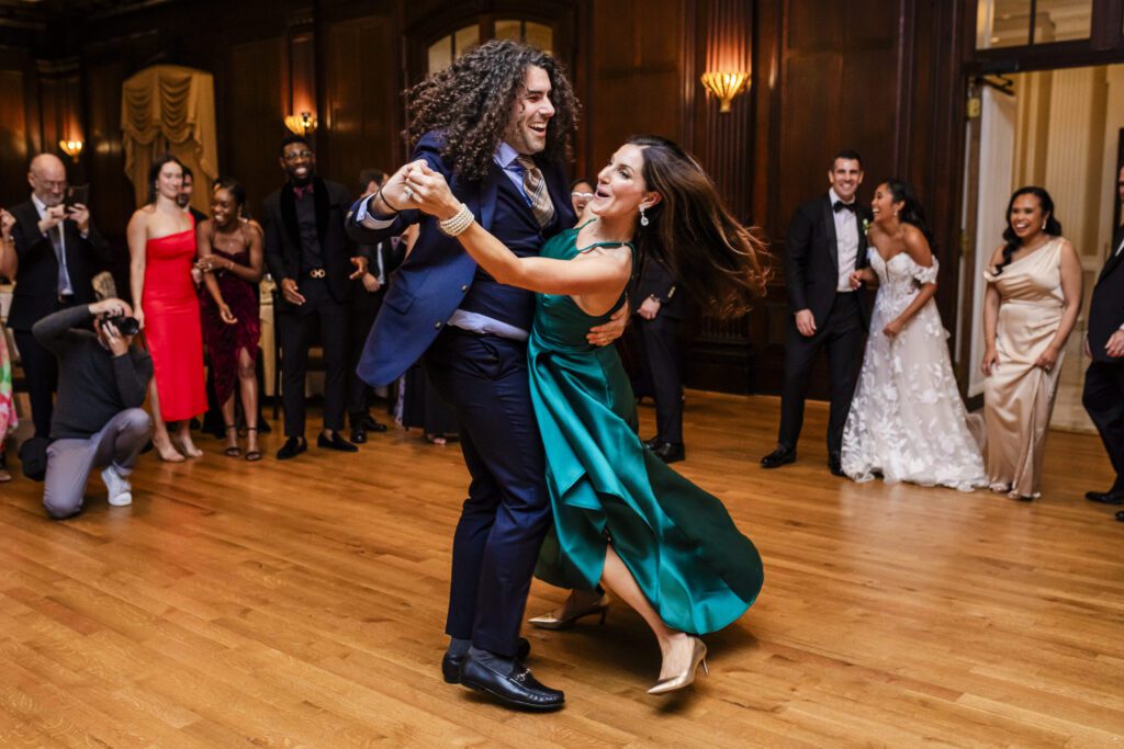 A young man with long curly hair twirls a woman in a green dress around the dance floor at the Union League while a bride & groom look on from the background laughing.